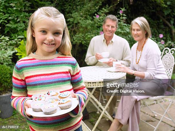 young girl in a garden holding a plate full of cup cakes with two adults sat behind her at a table, holding tea cups - cupcake teacup stockfoto's en -beelden