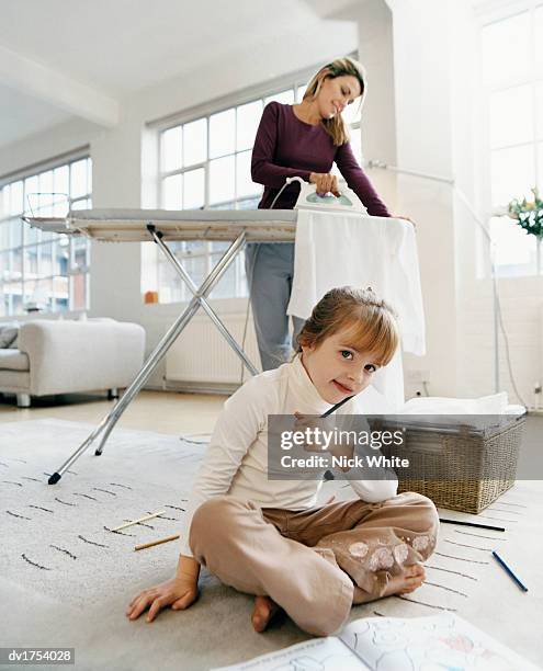 young girl sitting cross legged with a colouring in book on a rug with her mother ironing in the background - colouring fotografías e imágenes de stock