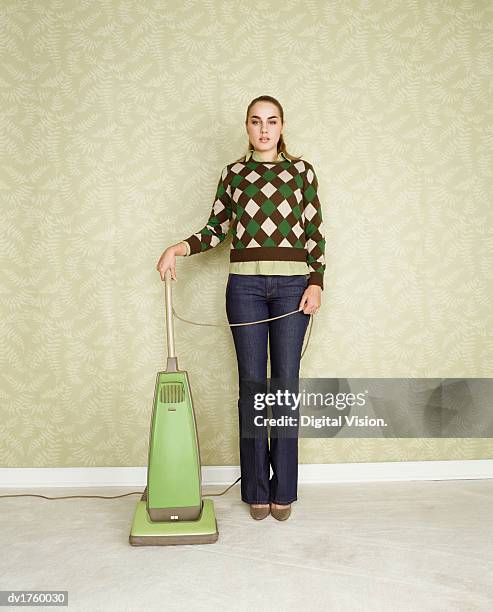 portrait of a woman standing next to a vacuum cleaner, against a wall decorated with green wallpaper - gestalt stock-fotos und bilder