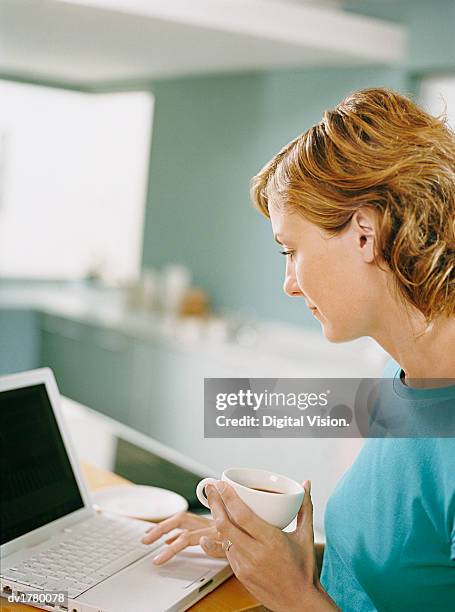 woman uses her laptop at a kitchen table, holding a cup of coffee - uses stock pictures, royalty-free photos & images