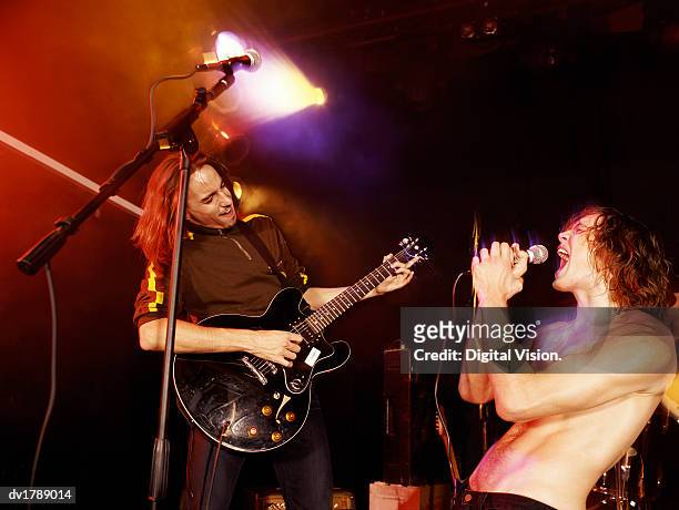 guitarist and topless male rock singer stand on a spot lit stage, singer screaming into a microphone - spot lit imagens e fotografias de stock