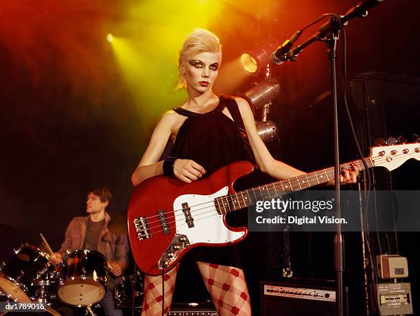 female rock band singer with exaggerated eye make-up and a blond quiff stands on stage with an electric guitar, drummer in the background - blonde female singers stock-fotos und bilder