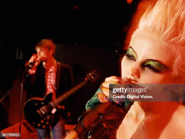 close-up of a female rock band singer with glittery eyeshadow and a blond quiff singing into a microphone on stage, male singer in the background - modern rock stock pictures, royalty-free photos & images