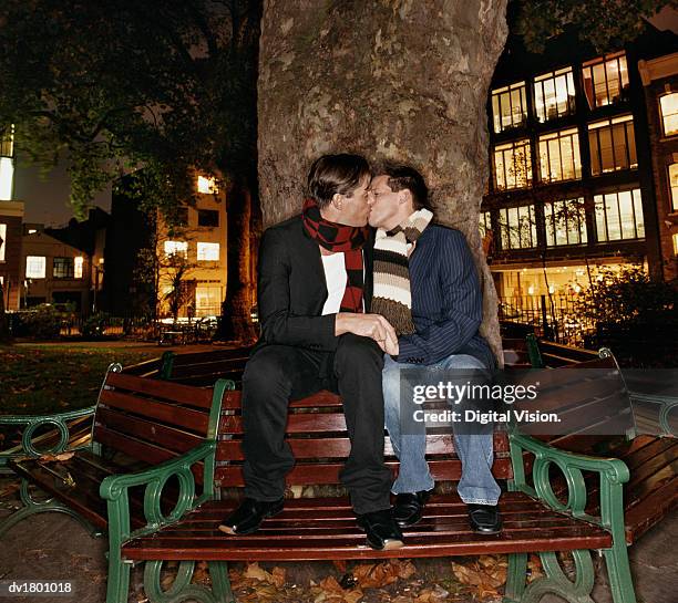 gay couple sit on a wooden bench in soho square kissing and holding hands - digital devices beside each other bildbanksfoton och bilder