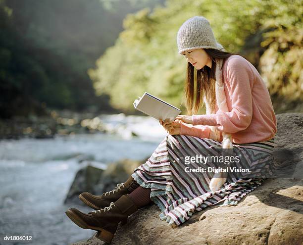 woman sat on a boulder by a river, reading a book - boulder rock stock-fotos und bilder