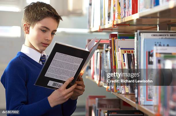 school boy stands next to a book shelf reading a book - book standing on end stock pictures, royalty-free photos & images