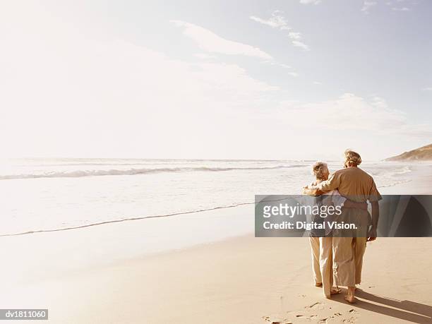 rear view of a senior couple walking on a beach with their arms around each other - high key foto e immagini stock