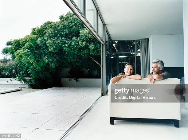 relaxed couple sitting on a sofa in their home with french windows open leading to a garden - porte d'accès à la terrasse photos et images de collection