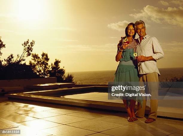 portrait of a glamorous senior couple holding cocktails by a swimming pool at sunset with the sea in the background - poolside glamour stock pictures, royalty-free photos & images