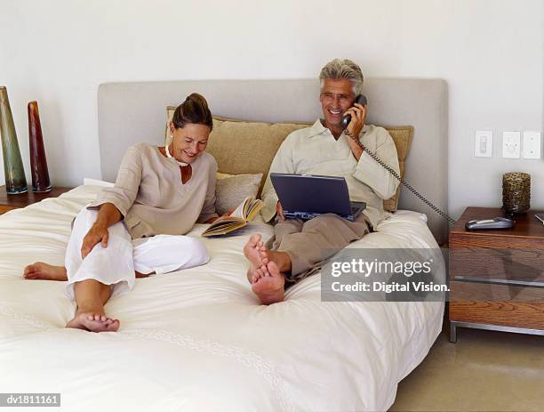 mature couple lie on their bed, woman reading a book and her husband using and laptop and talking on the phone - landline phone home stock pictures, royalty-free photos & images