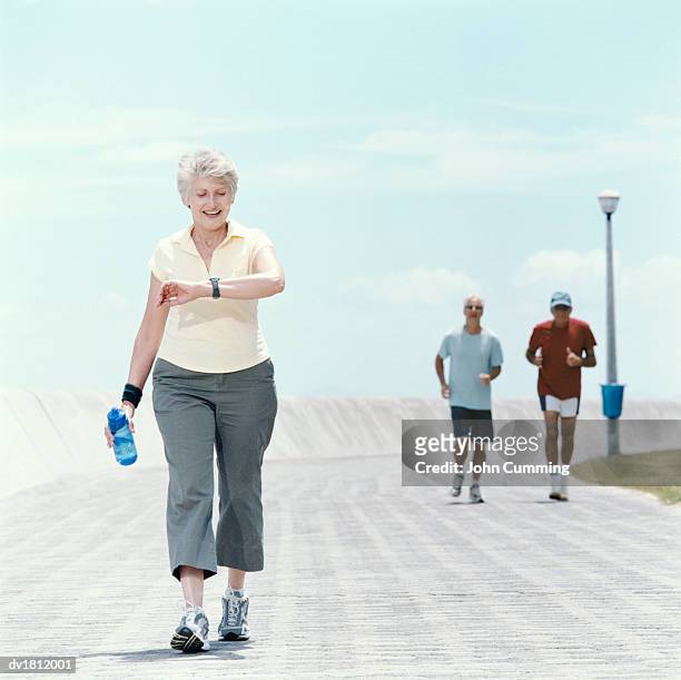 senior woman jogs on a path, checking her time on a watch - 3 old men jogging stock pictures, royalty-free photos & images