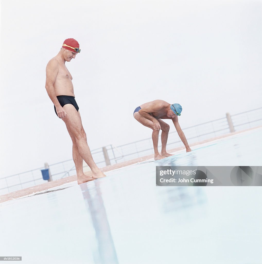 Two Senior Men In Swimming Trunks Stand at the Edge of a Swimming Pool, Testing the Temperature of the Water