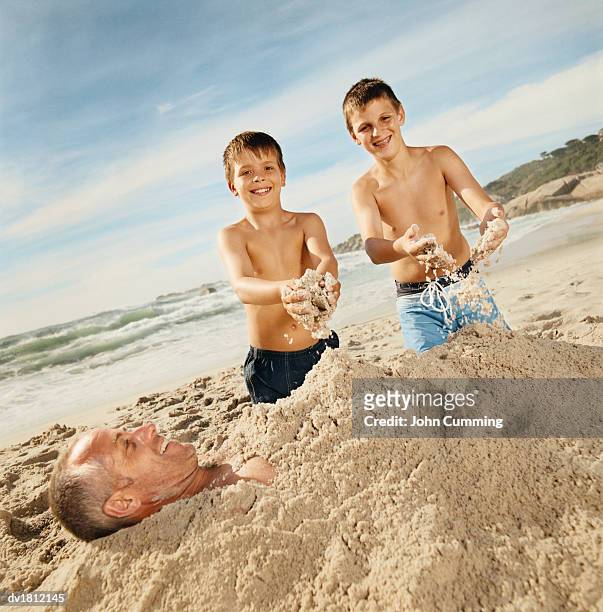 brothers on a beach burying their father in sand - buried in sand stock-fotos und bilder