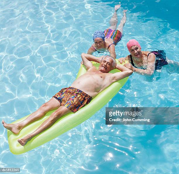 senior man in a swimming pool lying on an airbed which is being gripped by two senior women - man met een groep vrouwen stockfoto's en -beelden
