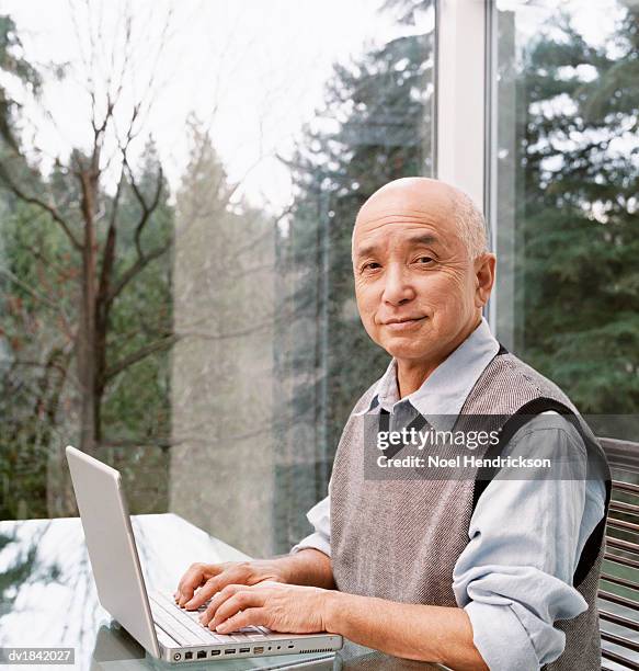 senior man sits at a table by a window using a laptop - silver surfer stock pictures, royalty-free photos & images