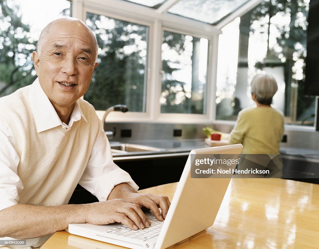 Senior Man Sits at a Table at Home Typing on a Laptop, His Wife in the Background