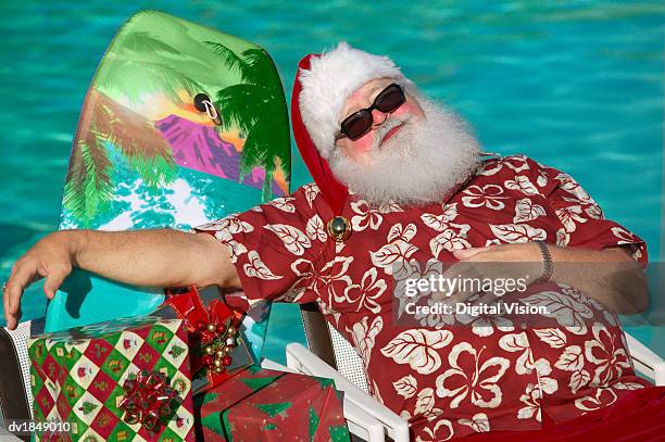 father christmas wearing a hawaiian shirt asleep in a chair by a swimming pool - santa swimming stock pictures, royalty-free photos & images