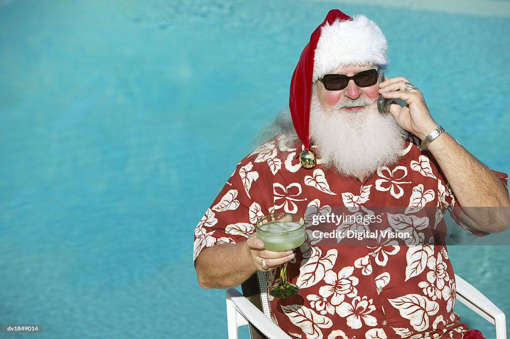 Father Christmas Wearing a Hawaiian Shirt Sitting by a Swimming Pool Holding a Mobile Phone and a Cocktail Glass