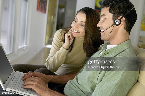 man sitting on a sofa wearing a headset using a laptop, watched by his partner - being watched stockfoto's en -beelden