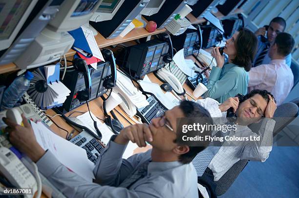 male stock trader sits at a desk with his colleagues making a phone call with his hand on his head in frustration - hand on head foto e immagini stock