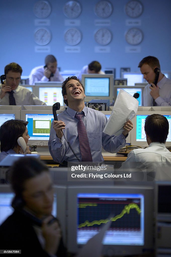 Male Stock trader Stands by His Desk in a Busy Office Holding a Phone Receiver and Printout and Looking up in Joy