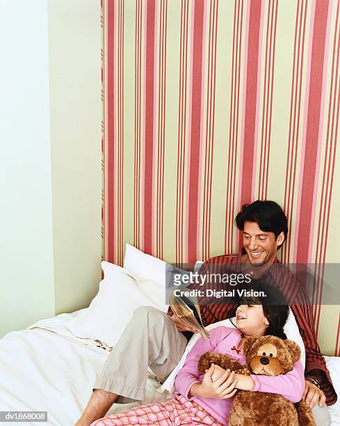 father and daughter lye on bed reading a book, girl holding a teddy bear - bedtime stories stock-fotos und bilder