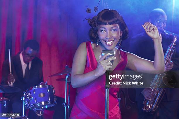 female jazz singer sings into a retro microphone, clicking her fingers, a saxophonist and drummer in the background - diva stock-fotos und bilder