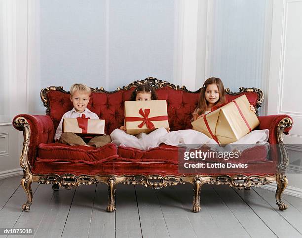 portrait of a three well-dressed young girls and boy sitting on an ornate velvet sofa holding wrapped gifts - ornate house furniture stock-fotos und bilder