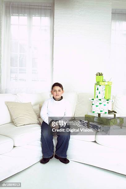 portrait of a young boy sitting patiently next to a stack of gifts on a white sofa - digital devices beside each other bildbanksfoton och bilder