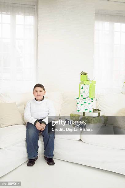 portrait of a young boy sitting patiently next to a stack of gifts on a white sofa - digital devices beside each other bildbanksfoton och bilder