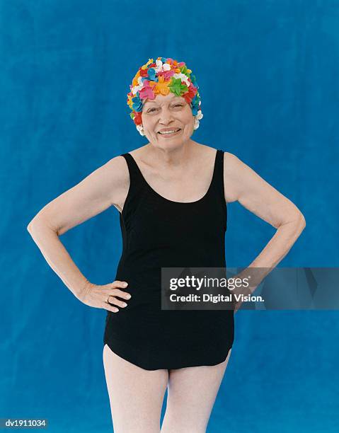 portrait of a senior woman standing in a swimming costume with her hands on her hips wearing a floral swimming cap - old woman in swimsuit stock pictures, royalty-free photos & images