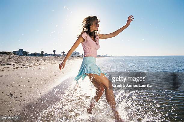 young, carefree woman in a miniskirt dances at the water's edge on the beach, splashing in the sea - lens flare young people dancing on beach stock pictures, royalty-free photos & images