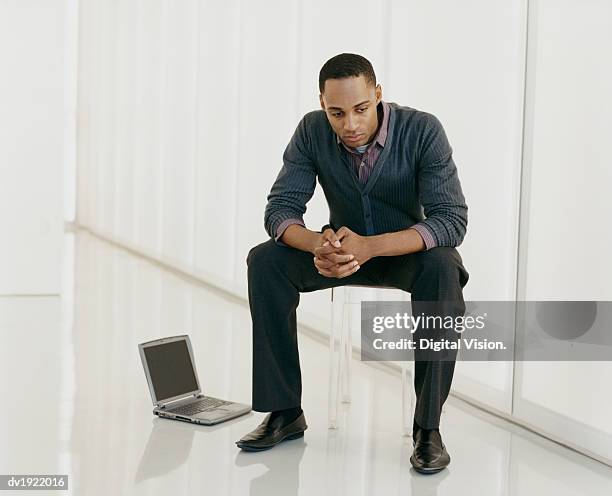 depressed looking man sitting on a transparent plastic chair, with a laptop on the floor - digital devices beside each other bildbanksfoton och bilder