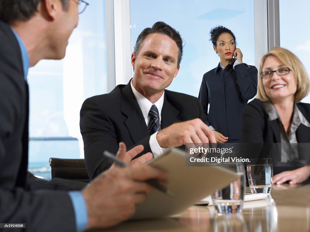 Business Executives in Discussion Sitting Around a Conference Room Table With a Businesswoman in the Background Using Her Mobile Phone