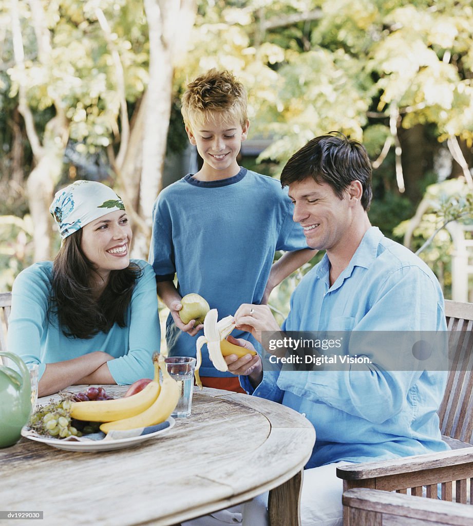 Father Peeling a Banana and Sitting at a Table in His Garden by His Son and Wife