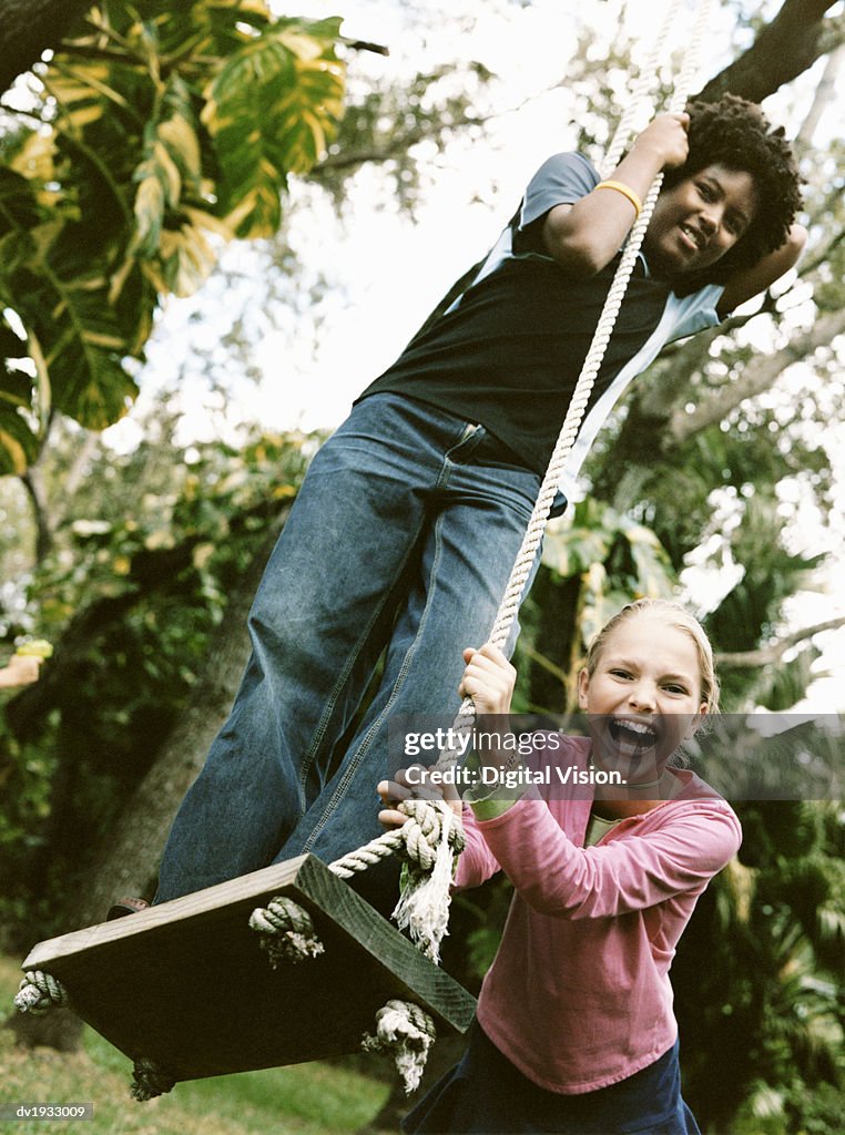 Young Girl Stands on Top of a Swing in a Garden, Being Pushed by Her Friend
