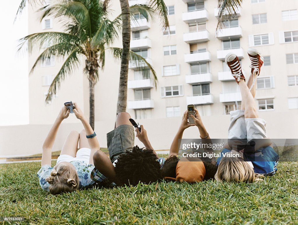 Rear View of Four Young Boys and Girls Lying on the Grass in a Line, Texting on Their Mobile Phones