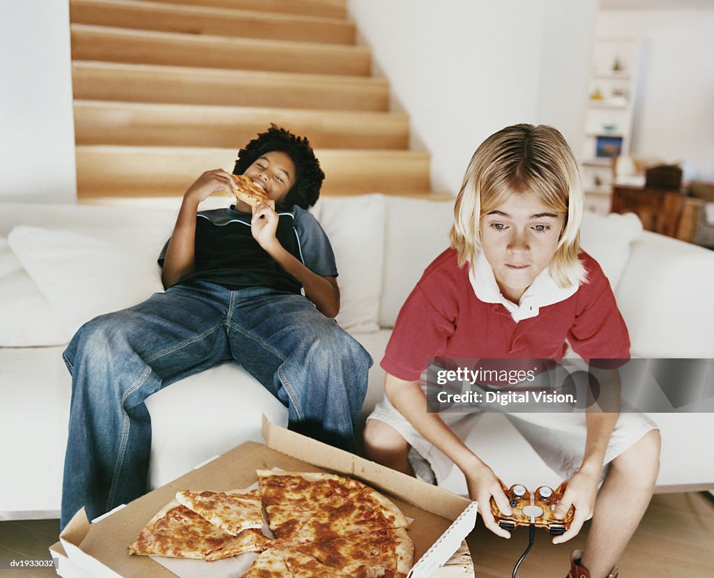 Two Boys Sitting on a Sofa, with One Boy Eating a Slice of Pizza and the Other Playing a Computer Game