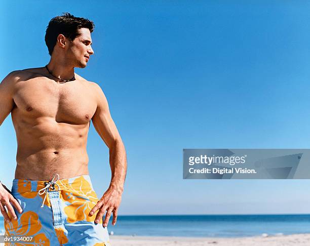 man standing on a beach with his hands on his hips and looking sideways - mens swimwear foto e immagini stock