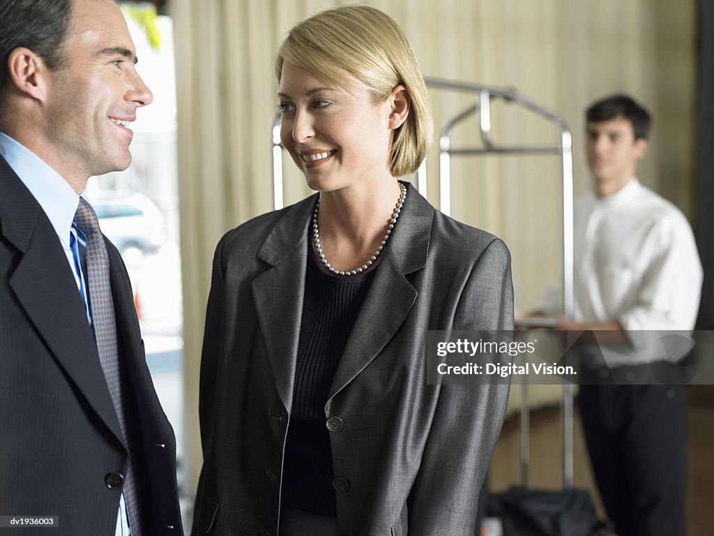 Businessman and Businesswoman Stand Talking to Each Other, a Hotel Porter in the Background