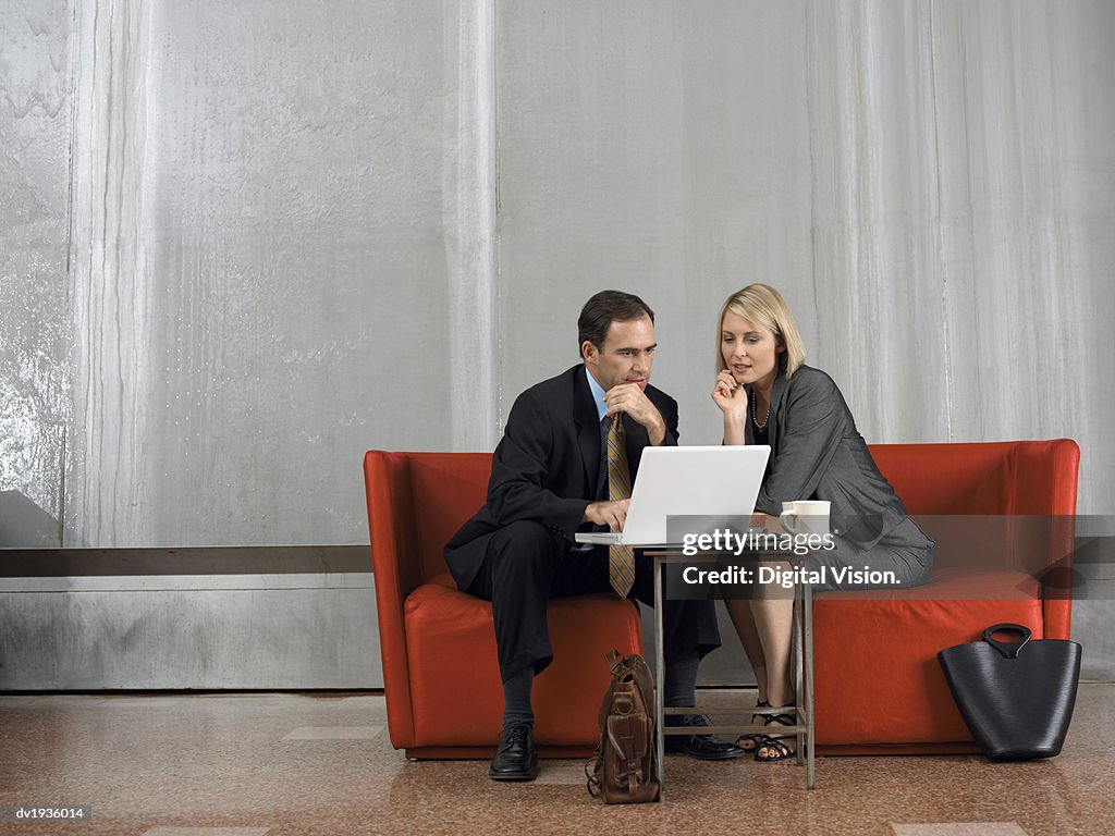 Businessman and Businesswoman Sit in Armchairs in a Lobby, Using a Laptop and Talking