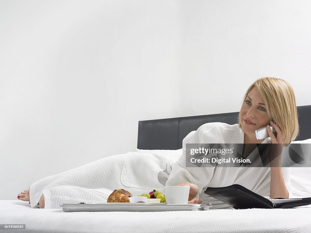 Woman Lies on a Bed in a Hotel Room With a Breakfast Tray and Using a Mobile Phone