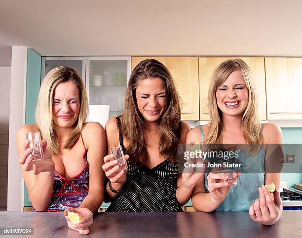 three teenage girls sit at a kitchen counter grimacing after drinking a shot of tequila - unangenehmer geschmack stock-fotos und bilder