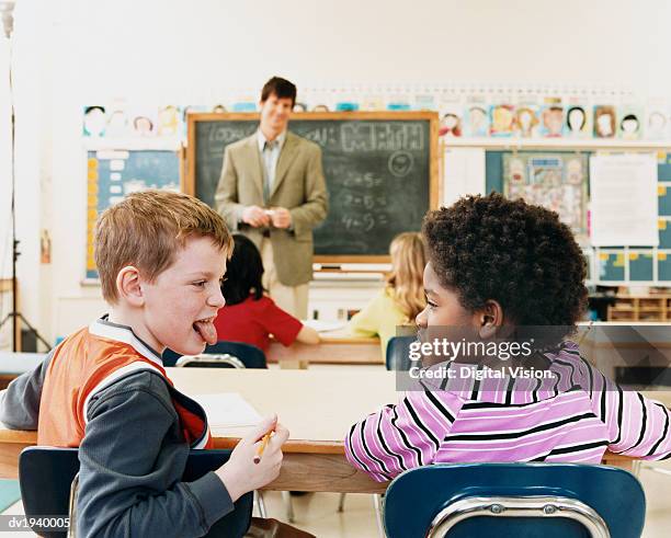 schoolboy and schoolgirl sitting in a classroom sticking their tongues out and a teacher in the background - naughty kids in classroom stock pictures, royalty-free photos & images