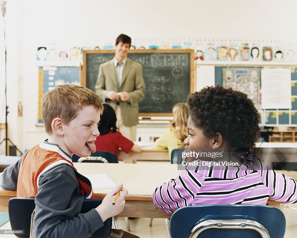 Schoolboy and Schoolgirl Sitting in a Classroom Sticking Their Tongues Out and a Teacher in the Background