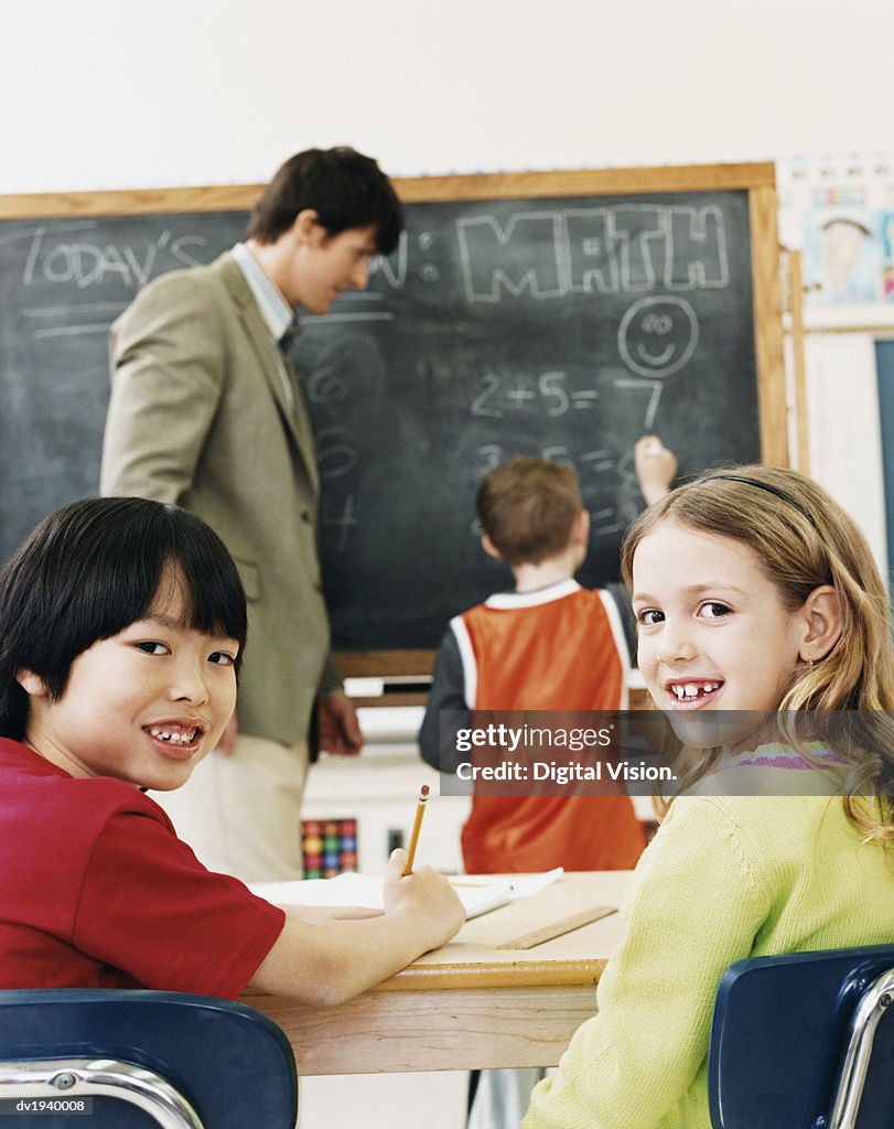 Primary Schoolboy and Schoolgirl Sitting at Desks and a Teacher and Schoolboy in the Background