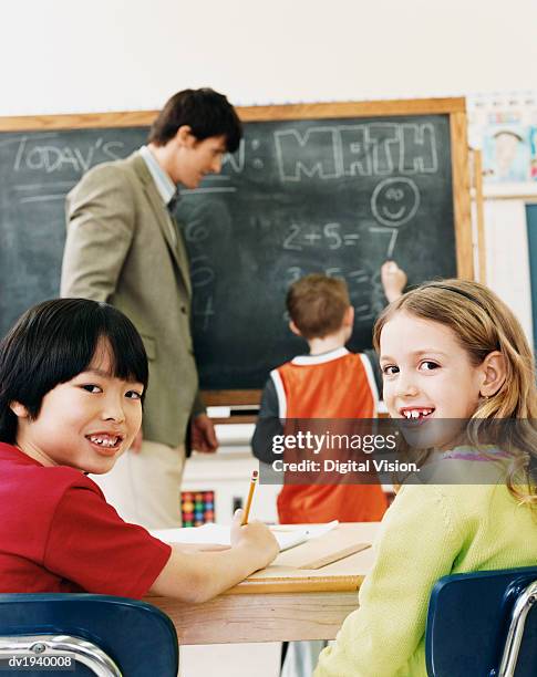 primary schoolboy and schoolgirl sitting at desks and a teacher and schoolboy in the background - boy looking over shoulder stock pictures, royalty-free photos & images