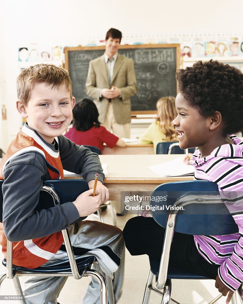 Primary Schoolboy and Schoolgirl Sitting Behind Desks in a Classroom and a Teacher in the Background