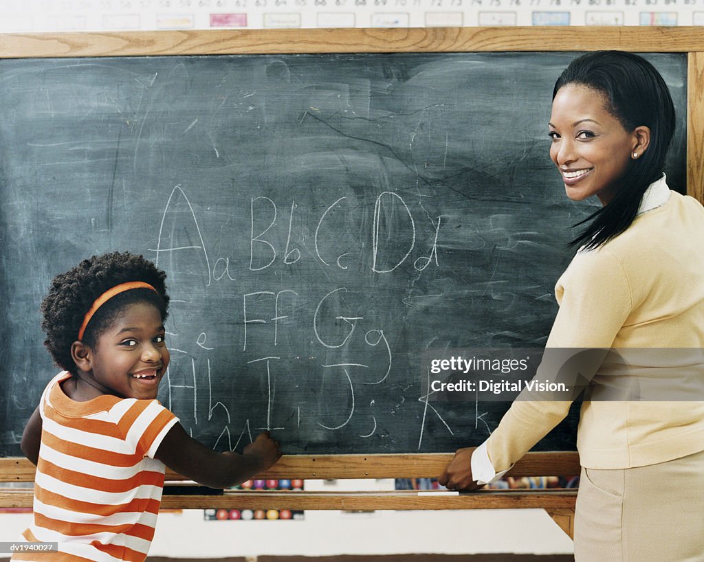 Primary Schoolgirl and Her Teacher Standing in Front of a Blackboard in a Classroom