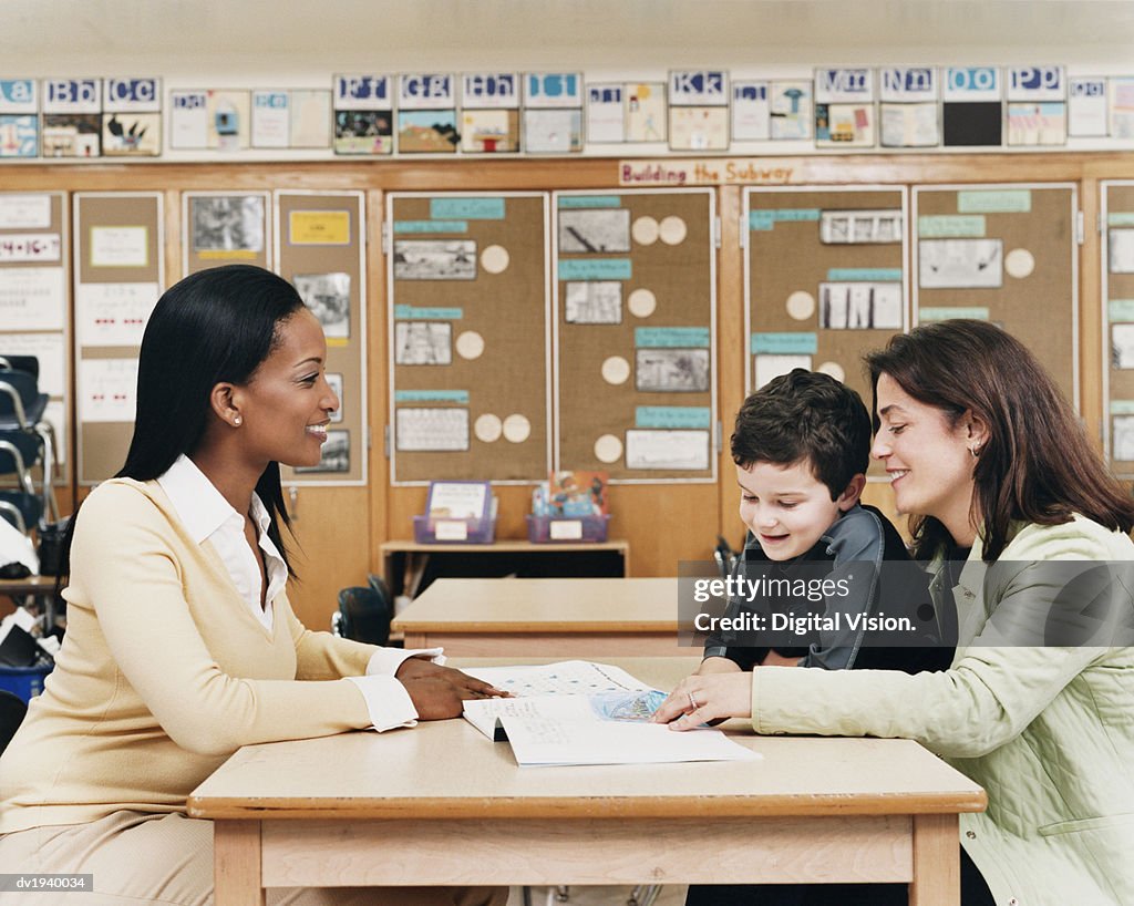 Teacher Sitting at a School Desk Showing a Book to a Parent and Her Son